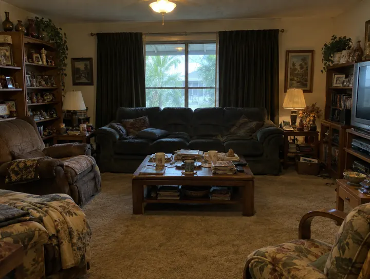 A living room before preparation, with heavy dark curtains, crowded shelves, mismatched furniture and dated carpet