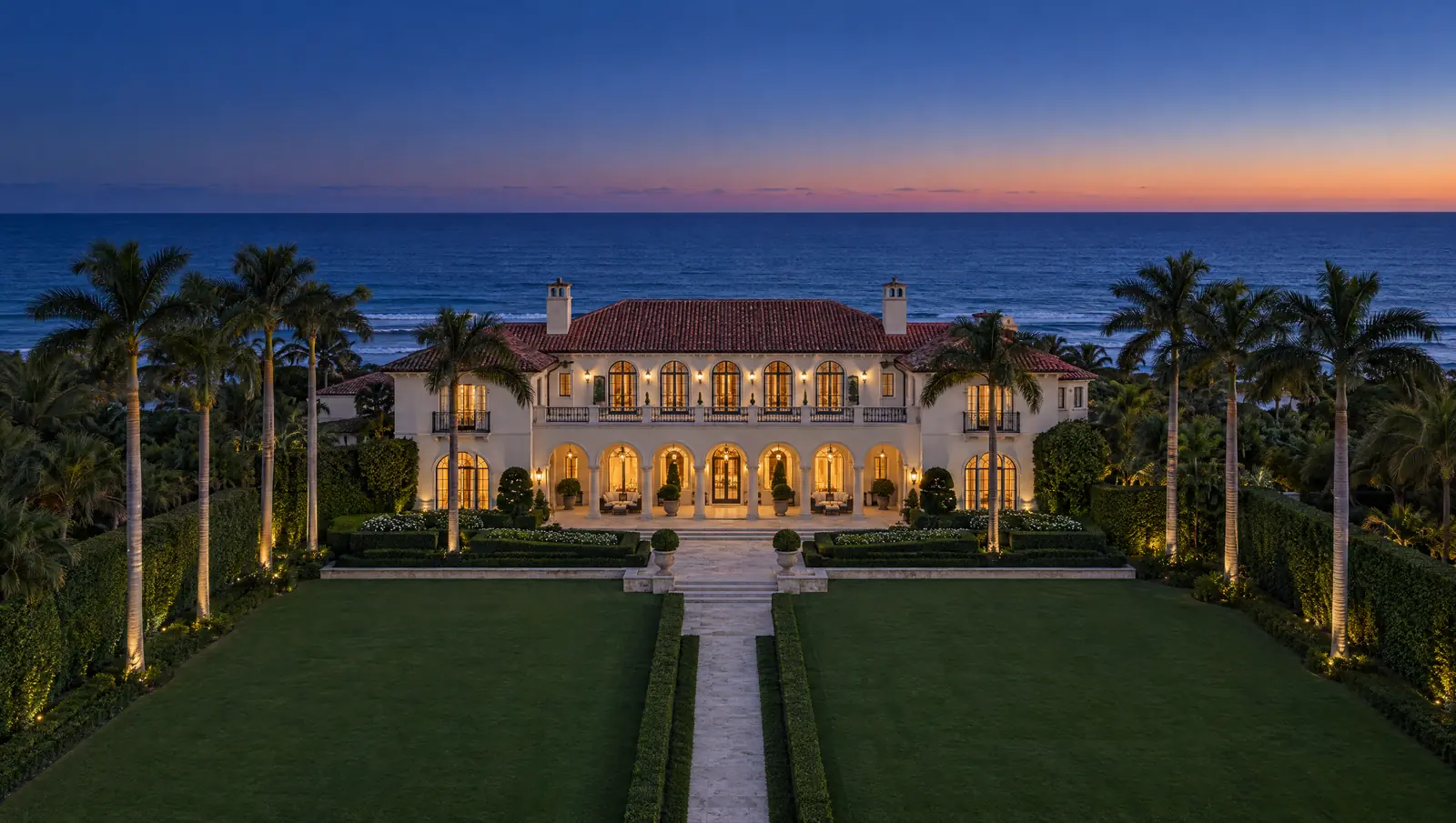 An aerial view at blue hour of a cream-coloured Mediterranean Revival estate on Palm Beach, its arched windows lit gold, a clipped lawn running to the Atlantic